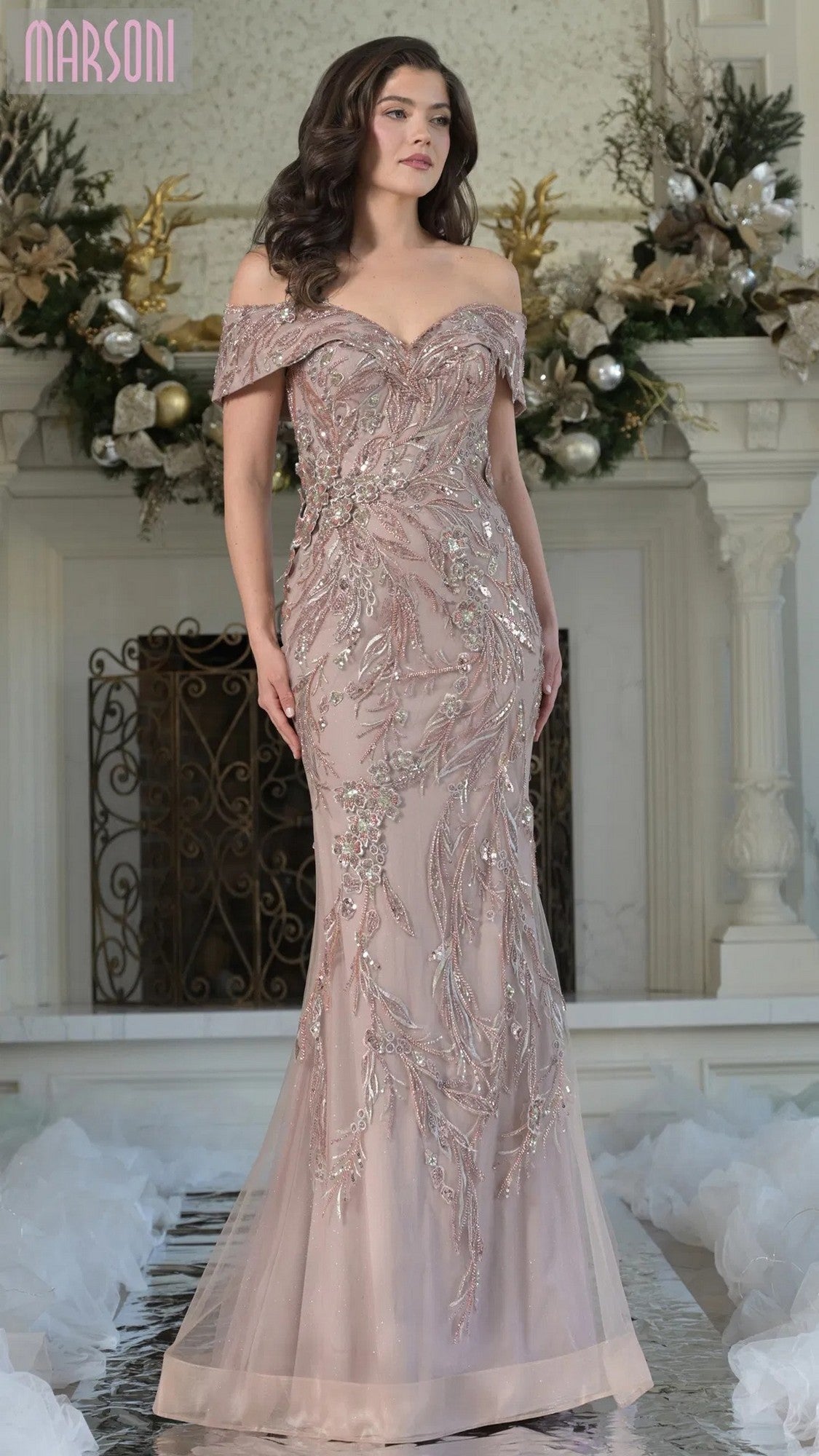 A woman stands indoors wearing the Marsoni MV1372 Long Formal Dress in taupe, an off-shoulder mermaid gown highlighted by intricate beaded embroidery. Floral decorations adorn the background.
